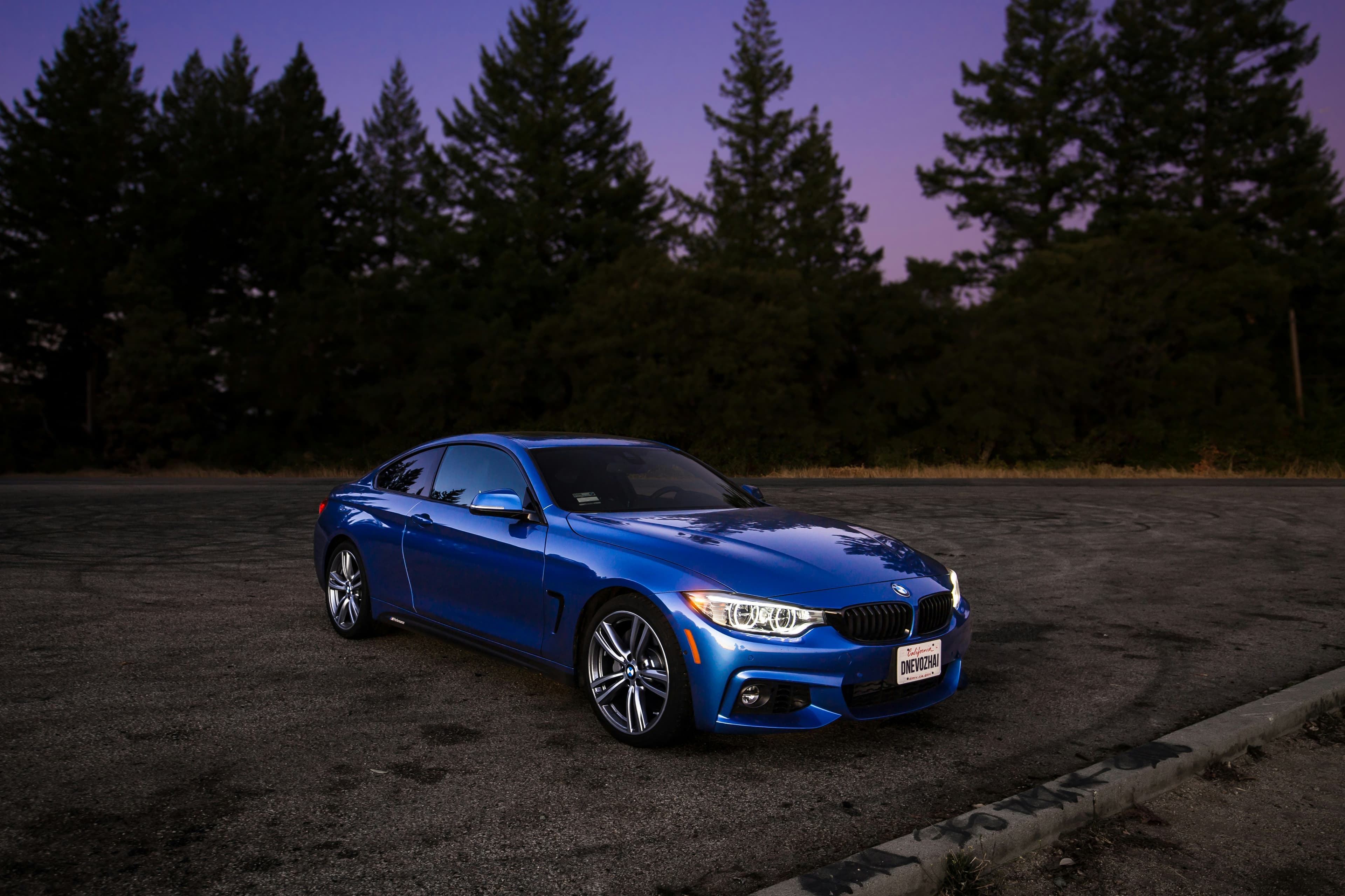 Blue BMW sedan parked on asphalt, surrounded by trees at dusk, with headlights on.