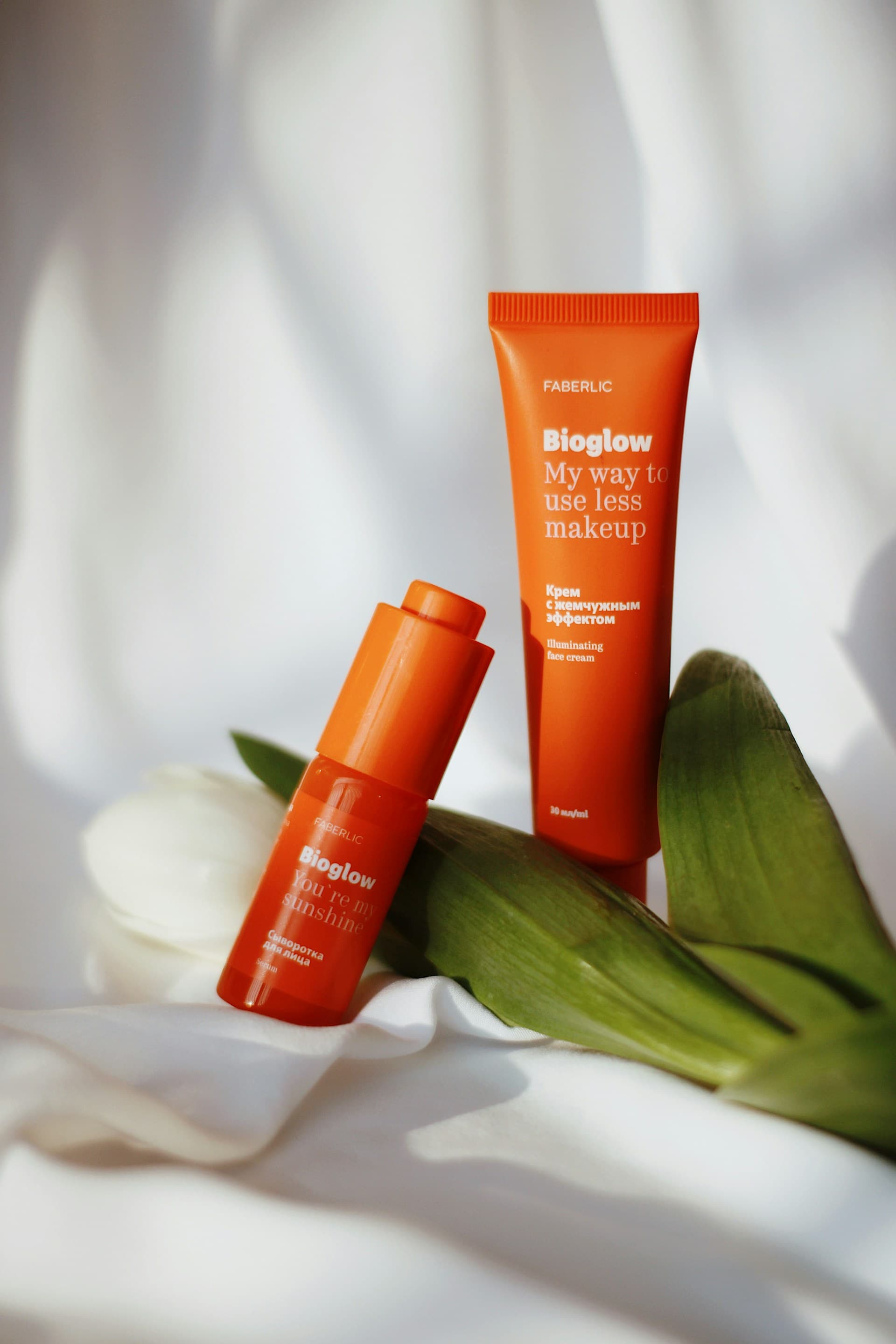 Orange Bioglow skincare products by Faberlic, including serum and cream, with green leaves and a white tulip.